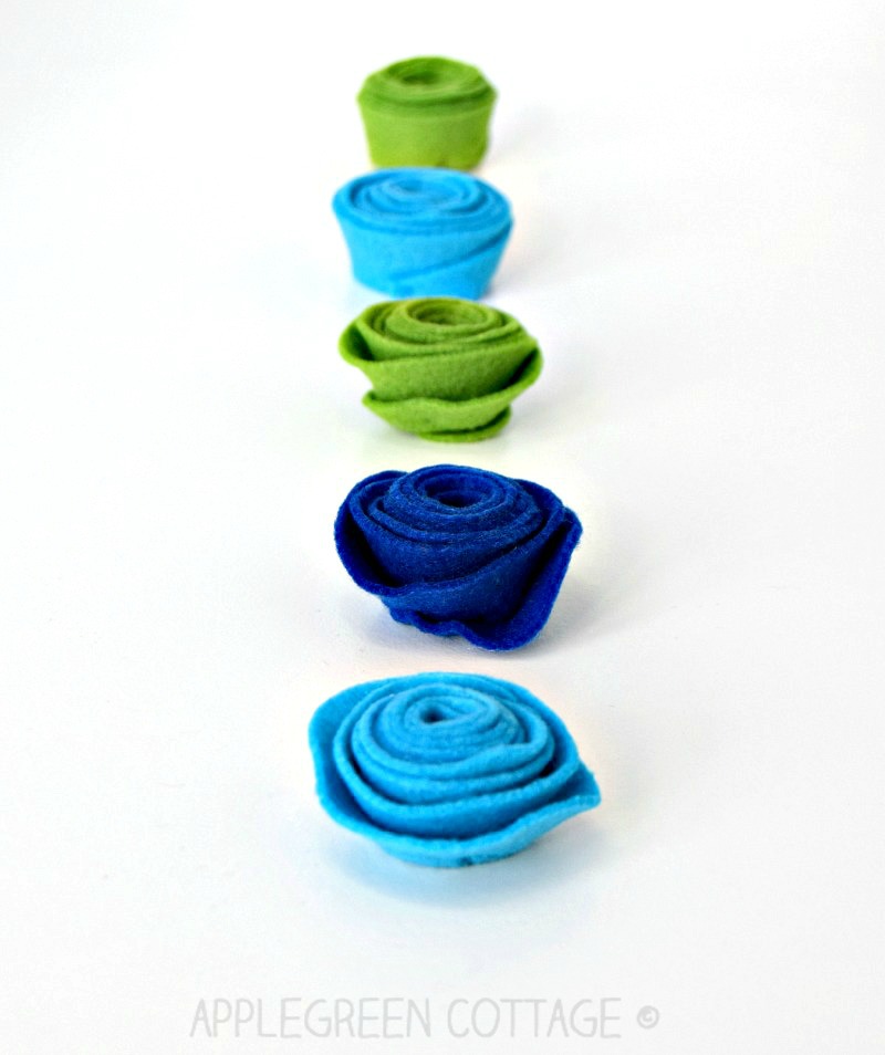 finished rolled flowers from green, aqua and blue felt arranged in a line