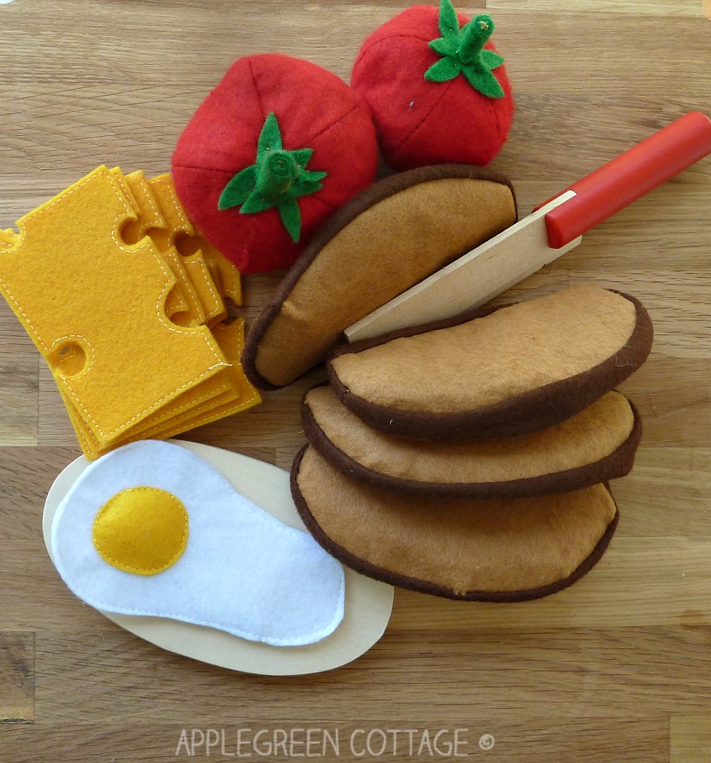 a set of diy felt play food items on a wooden table: a tomato, an egg, a slice of cheese and pieces of french loaf bread made out of felt