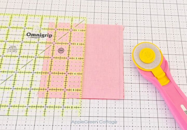 Quilter using a rotary cutter to trim the edges of a pink fabric square on a gridded cutting mat.