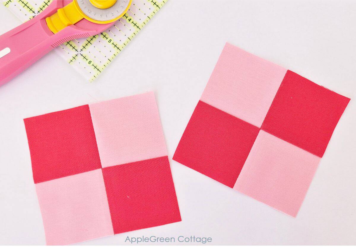 Two completed pink and red four-patch quilt blocks placed on a cutting mat, with a rotary cutter beside them.