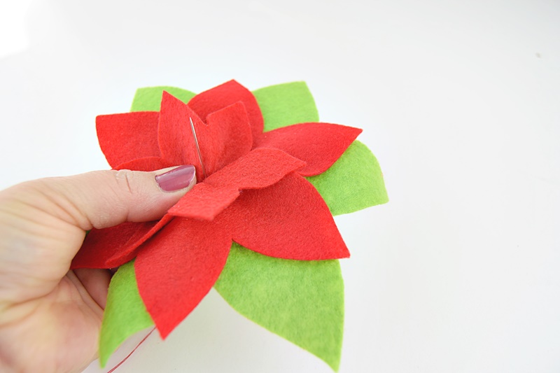 a hand holding red felt leaves and a sewing needle, assembling a diy poinsettia ornament