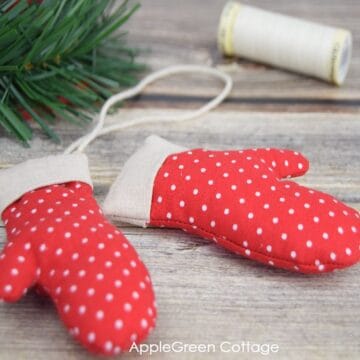 two small fabric mittens sewn with polka dot red fabric and placed on a wooden surface next to a christmas tree and a spool of gรผtermann cotton thread