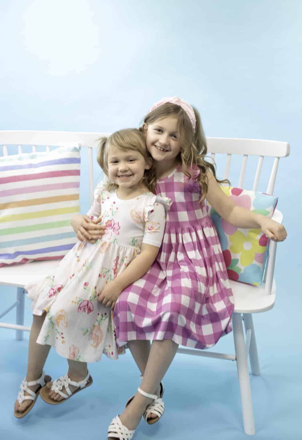 two girls smiling and sitting on a bench wearing homemade dresses in cute fabric prints