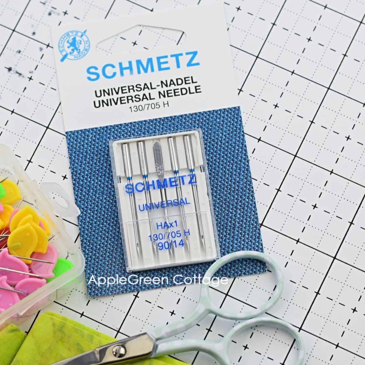 a box of schmetz universal sewing machine needles placed next to snipping scissors and flat pins for sewing