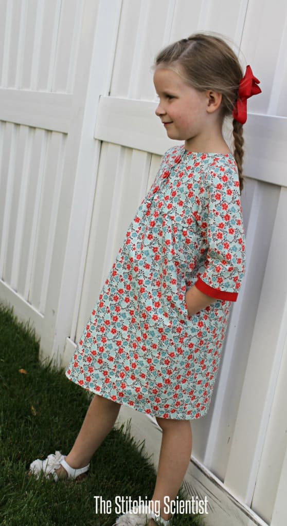 a girl in a homemade floral empire dress with a red ribbon bow, standing next to a white wooden fence