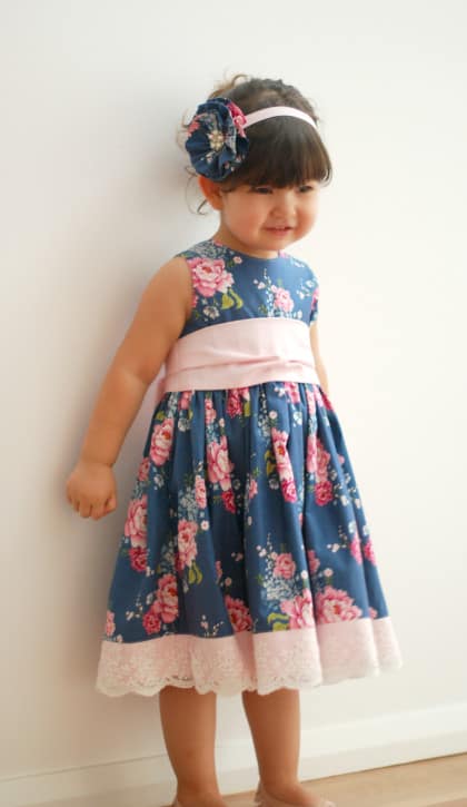 a girl in a blue floral homemade dress with white lace trim standing next to a white wall