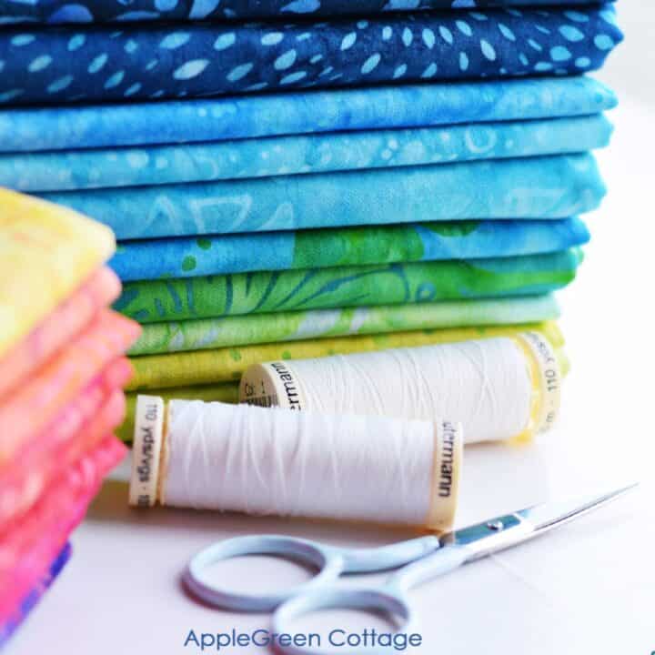 fabric stacked on a table and arranged by color next to small scissors and spools of thread