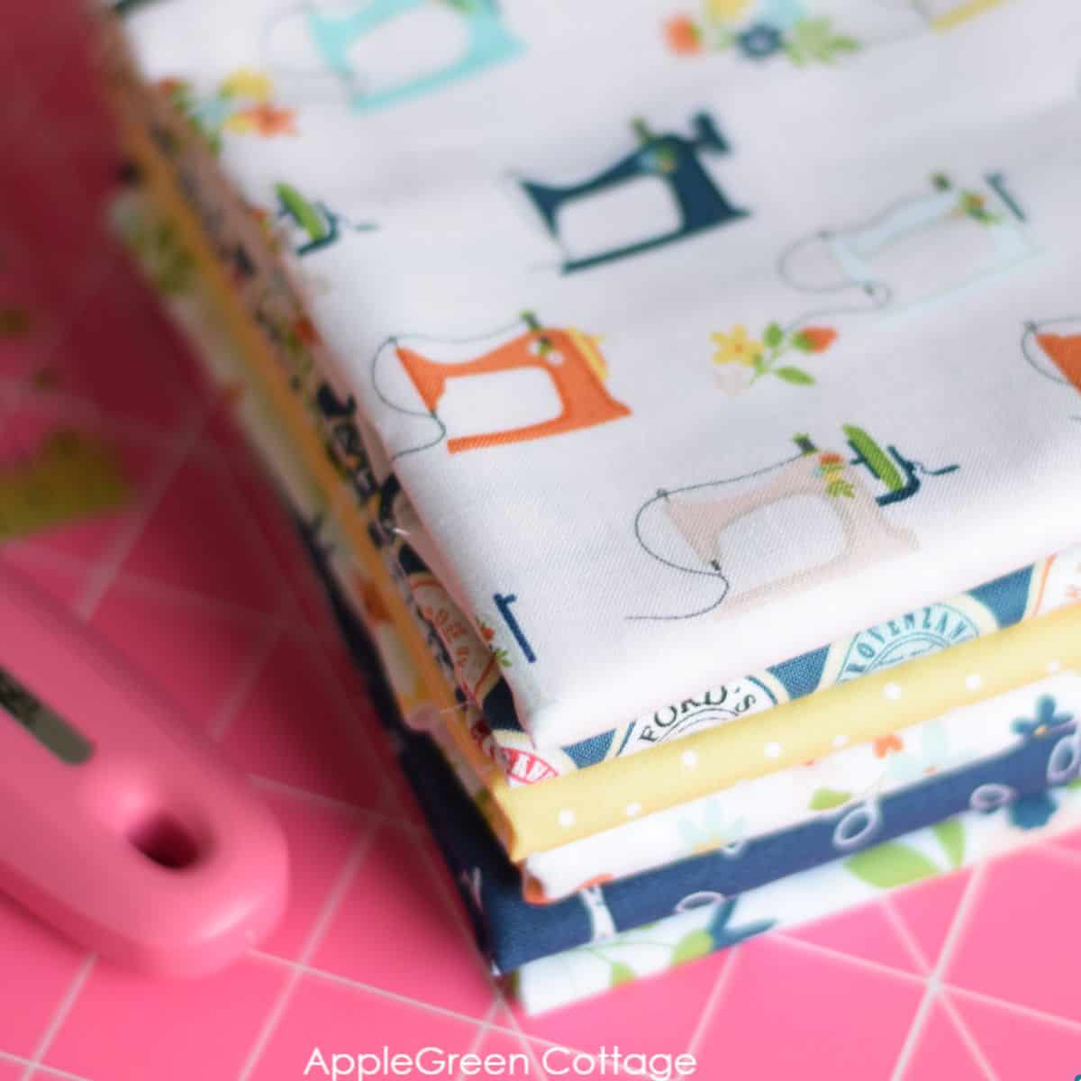 a stack of quilting cotton fabric collection on a pink cutting mat, next to a pink rotary cutter