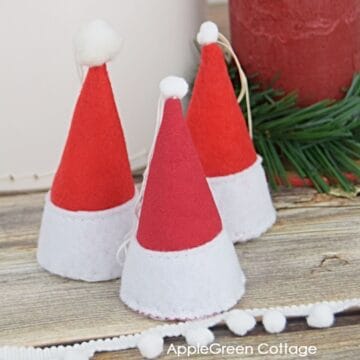 three finished handmade Santa hat ornaments in red and white felt and fabric with a candle and a spruce twig in the background