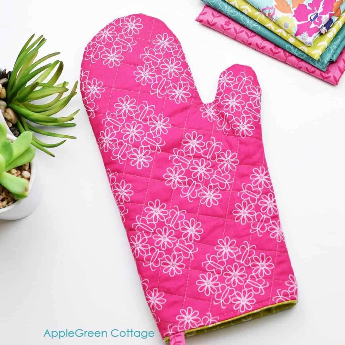a finished pink quilted oven mitt with floral print on a table next to green flowers