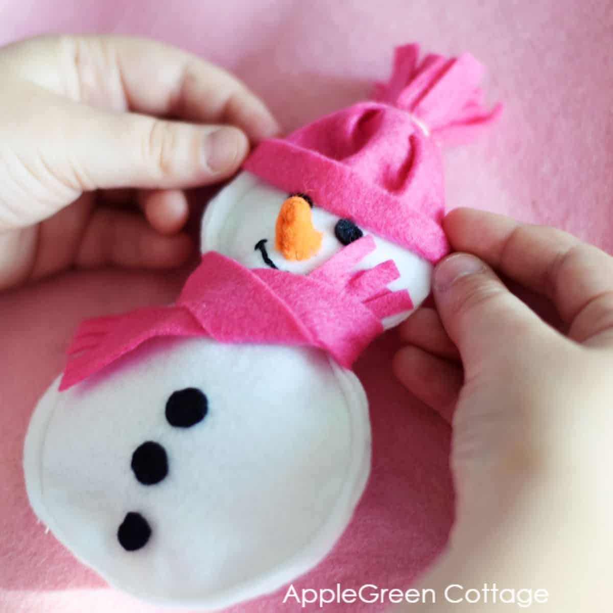 two hands putting on a pink hat on a snowman ornament