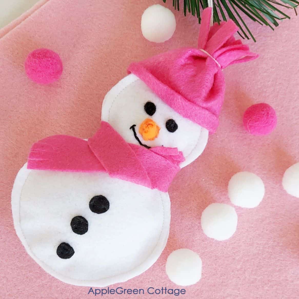 a white and pink felt snowman ornament on a light pink background next to small pompom decorations