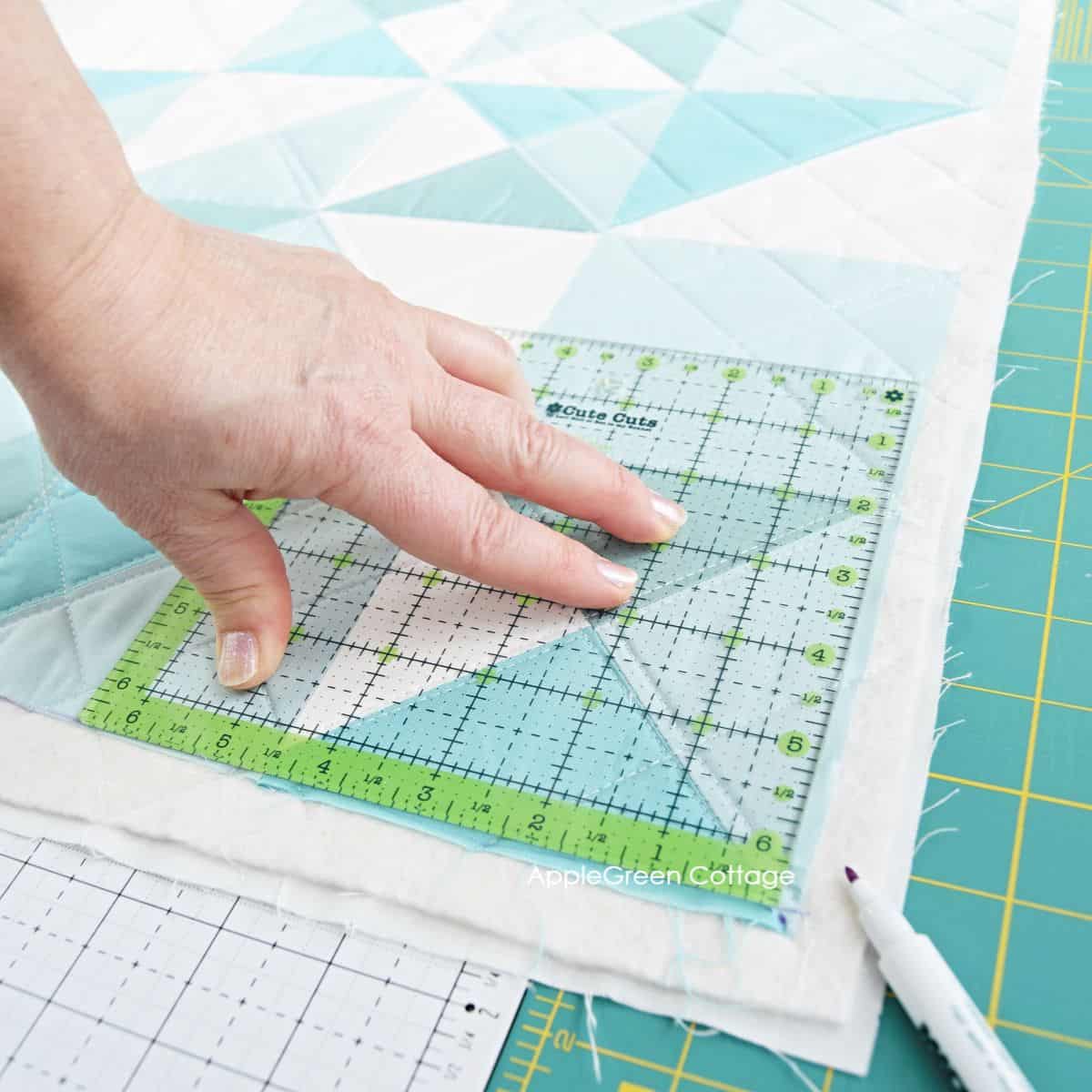 a hand holding a quilting ruler in place, indicating a diagonal direction in the corner of an aqua quilt that is about to be squared up