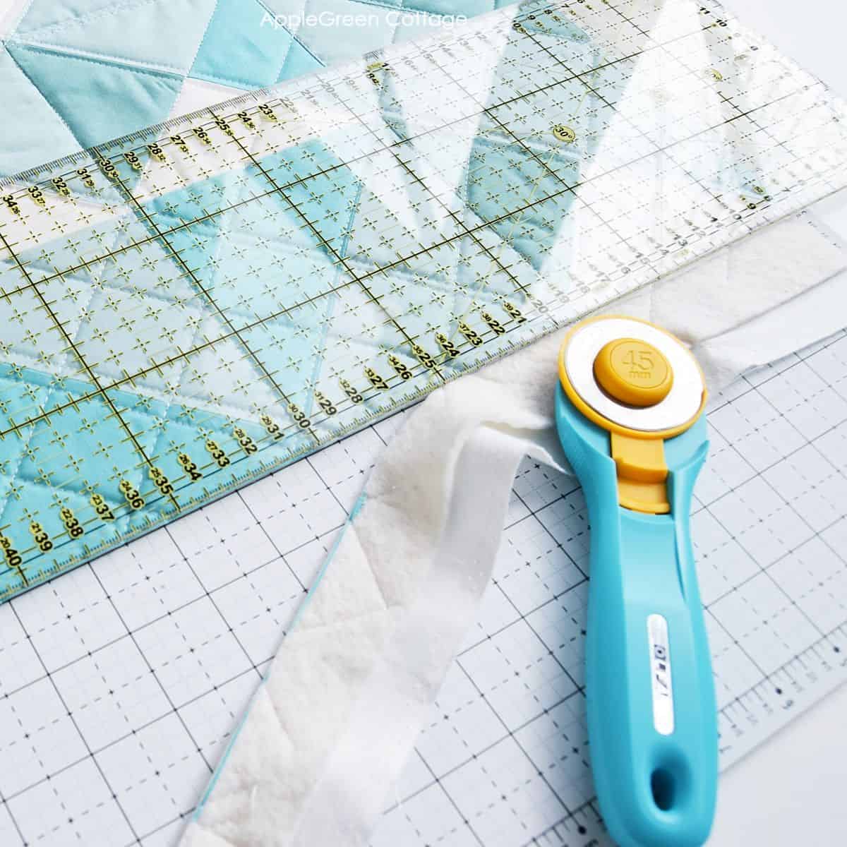 aqua rotary cutter cutting along a quilt edge using a large quilting ruler, placed next to a piece of already cut excess batting and fabric 