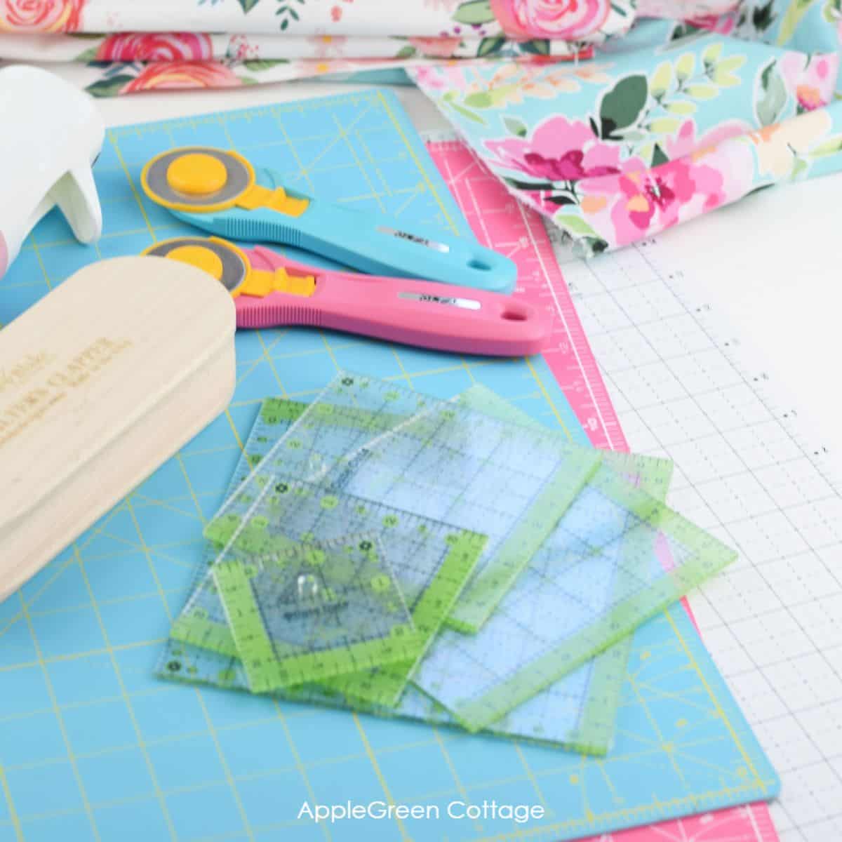 pink rotary cutter and an aqua olfa rotary cutter placed on a white cutting mat next to a set of quiltign rulers and beautiful floral quilting cotton print
