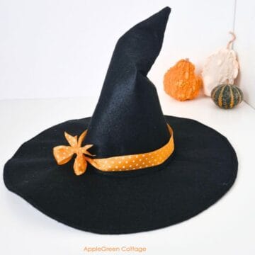 a black witch hat sewn from black felt with a wide brim, an orange polka dot ribbon and a crooked hat top, placed next to three pumpkins