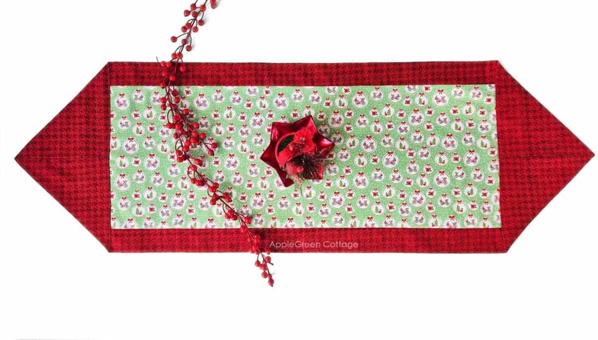 a finished fabric table runner sewn with two fabric pieces in Christmas prints, with a red glossy star-shaped plate on it and 2 stems of holly red berries