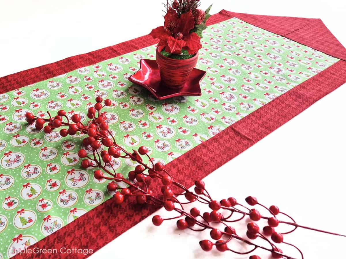 a finished fabric table runner sewn with two fabric pieces in Christmas prints, with a red glossy star-shaped  plate on it and 2 stems of holly red berries 