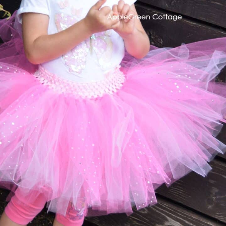 a small girl standing next to a wooden fence, wearing a handmade pink tulle tutu skirt, a pair of pink leggins and a lovely white t-shirt