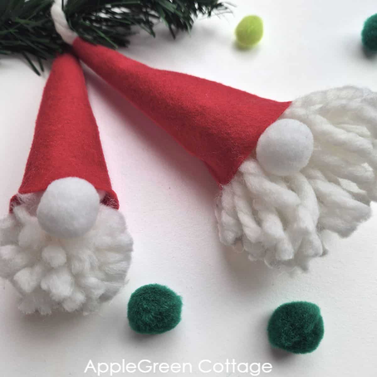 two homemade pompom gnome ornaments with red felt hats and white yarn heads with holiday decorations in green