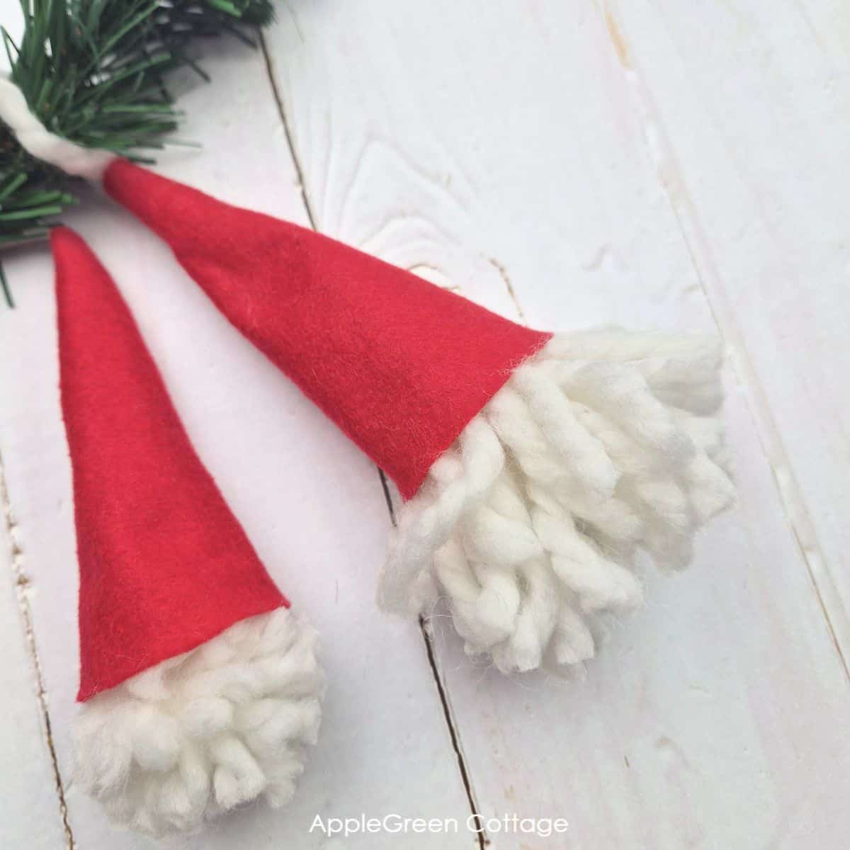 two simple pompom gnome ornaments without noses with red felt hats and white yarn heads hung on a small spruce twig