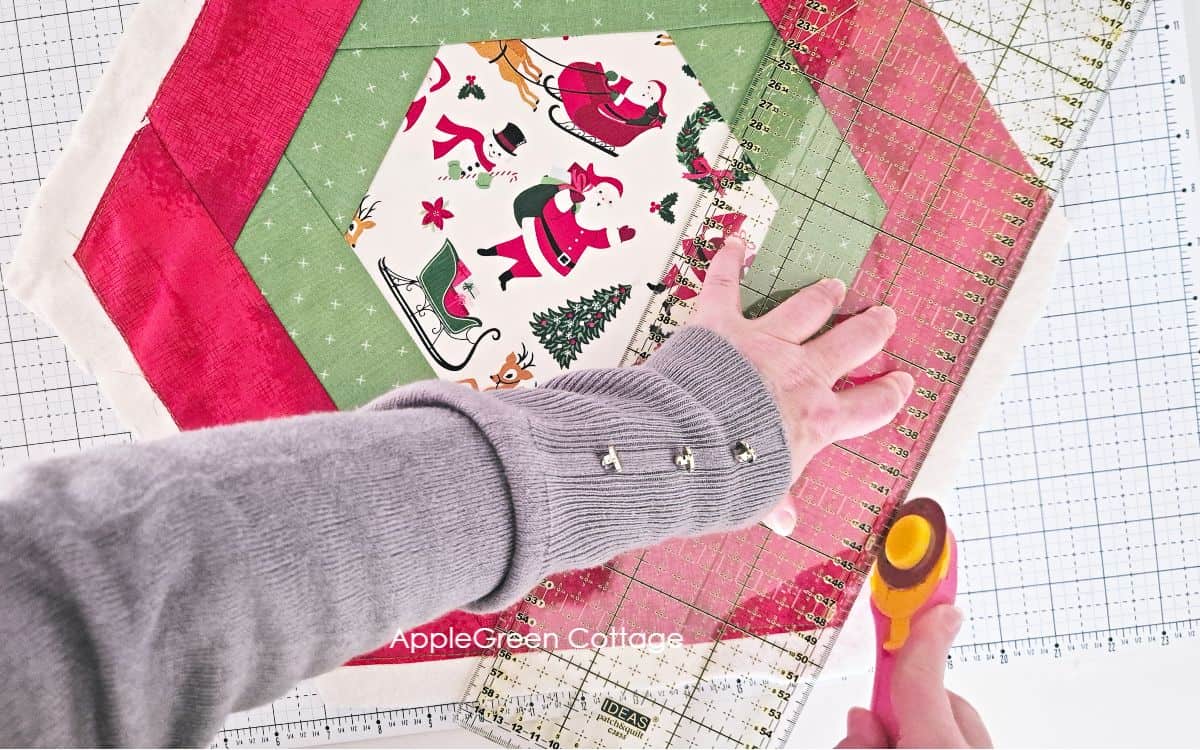 a pieced red hexagon table runner placed on a cutting mat and partially covered by a hand holding a pink rotary cutter next to a long quilting ruler