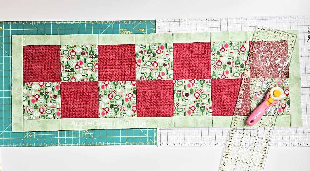 a pieced quilt top covered by a square quilting ruler, placed on a cuttinng mat next to a pink olfa rotary cutter