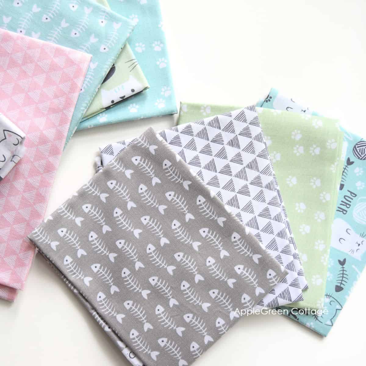 A stack of colorful fat quarters featuring playful designs, including geometric patterns, cat prints, and fishbone motifs in soft pastel colors like pink, mint green, and gray. Perfect for sewing and crafting projects.