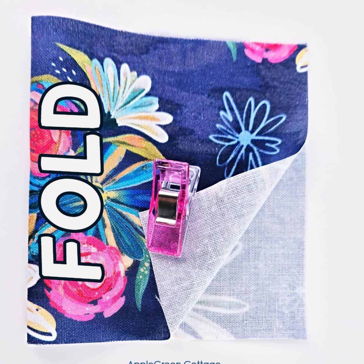 A floral fabric square folded and clipped, labeled 'FOLD' with a bright pink sewing clip securing the layers.