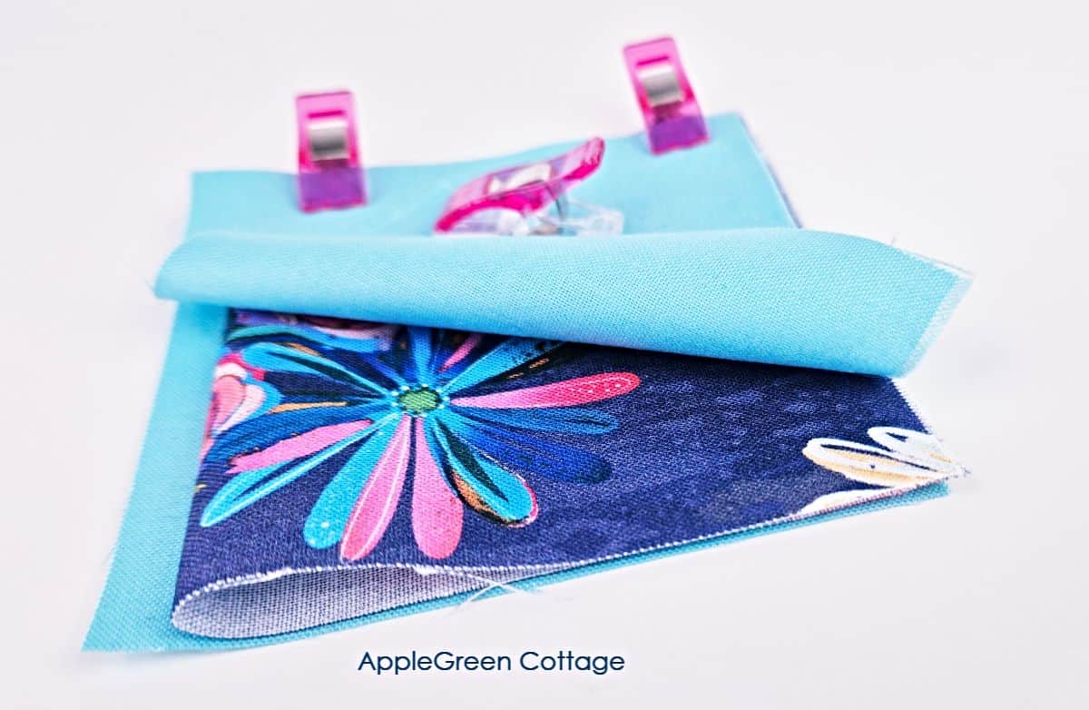 Layers of floral and blue fabric folded together, clipped with pink sewing clips, demonstrating alignment for sewing.