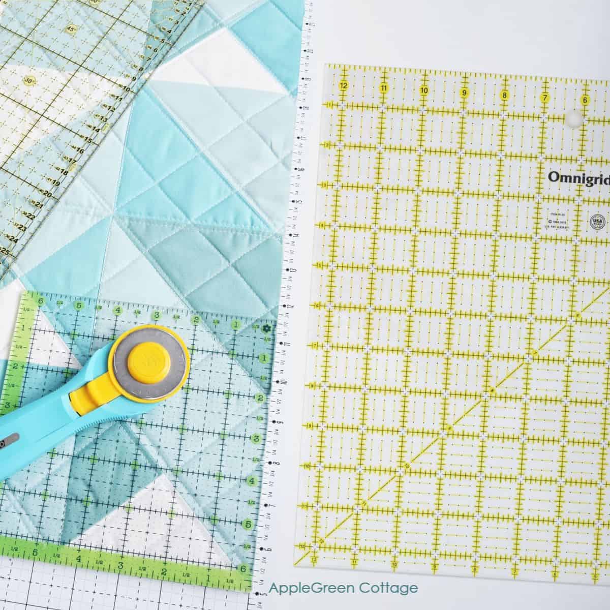 A collection of quilting rulers, including Omnigrid and square rulers, alongside a rotary cutter and a quilted aqua and white fabric design. Perfect tools for accurate quilting and precise cuts.
