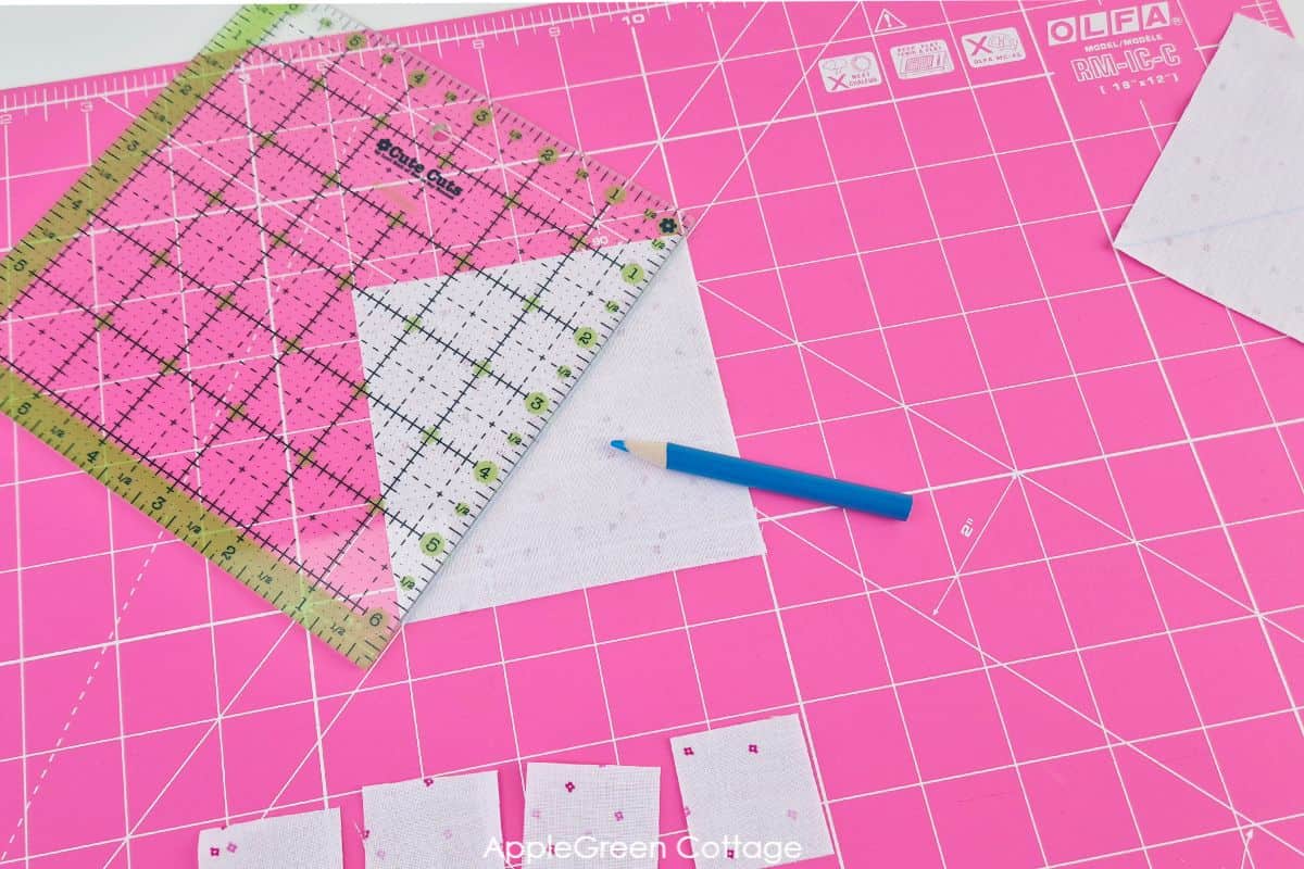 Marking diagonal lines on white fabric squares using a quilting ruler and blue marking pencil on a pink cutting mat.