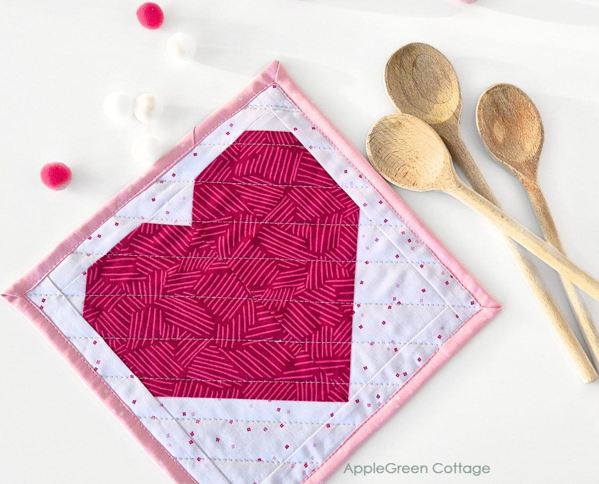 A pink quilted potholder with a bold heart design, surrounded by wooden spoons for a cozy kitchen theme.