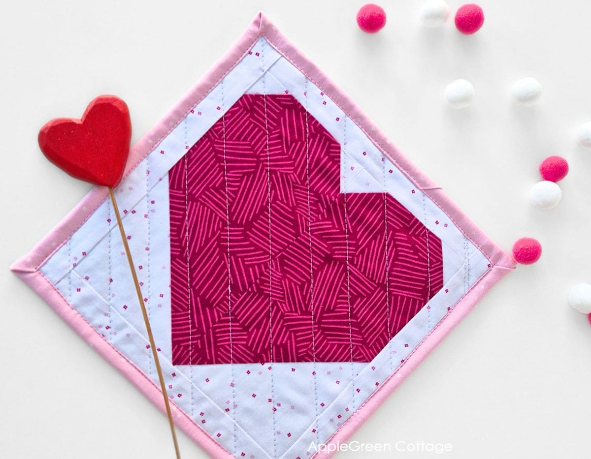 A completed quilted pink heart potholder, styled with Valentine's decor elements like pom-poms and hearts.