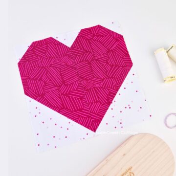 A heart quilt block pieced with vibrant pinks and a light white blender background, next to a quilter's clapper and embroidery scissors.