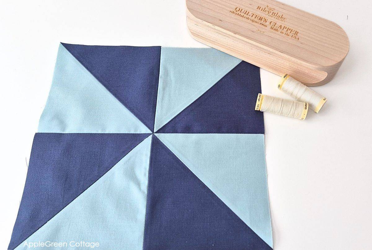 Finished Pinwheel Quilt Block with Quilter’s Clapper – A neatly sewn pinwheel quilt block in navy and light blue fabric, with a quilter’s clapper and spools of thread beside it.