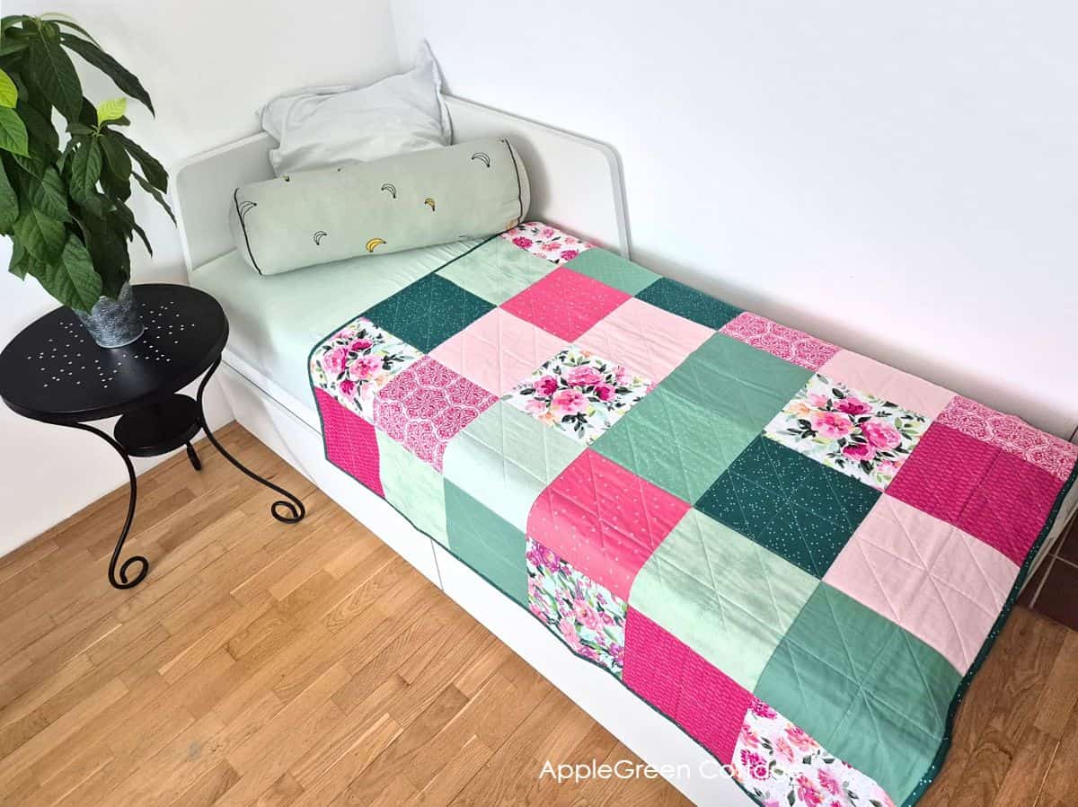 Pink and green patchwork quilt displayed on white daybed with decorative pillow, featuring floral fabric squares and geometric design.