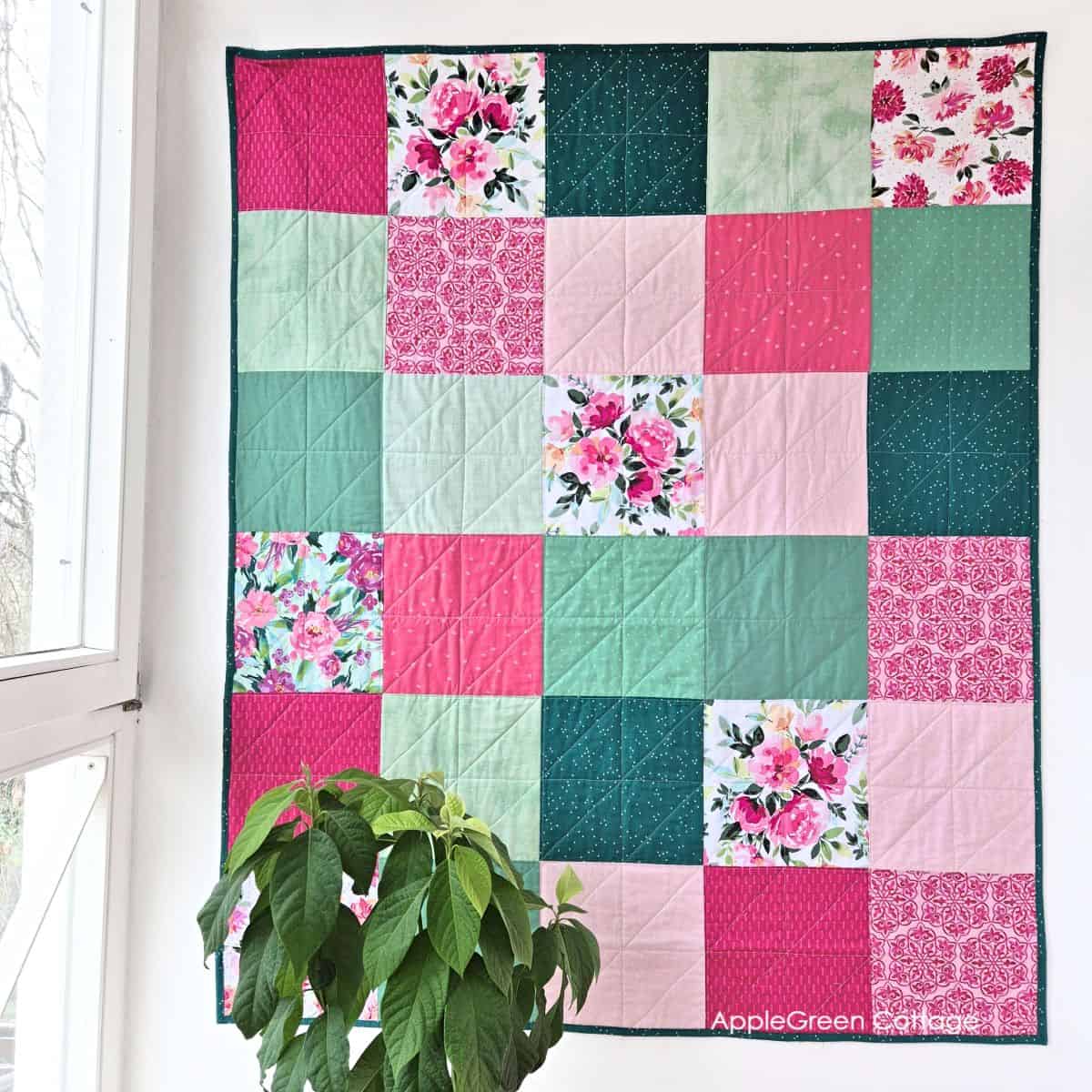 Modern floral patchwork quilt hanging by window with square pattern in pink, green, and teal colors with consistent quilting lines.