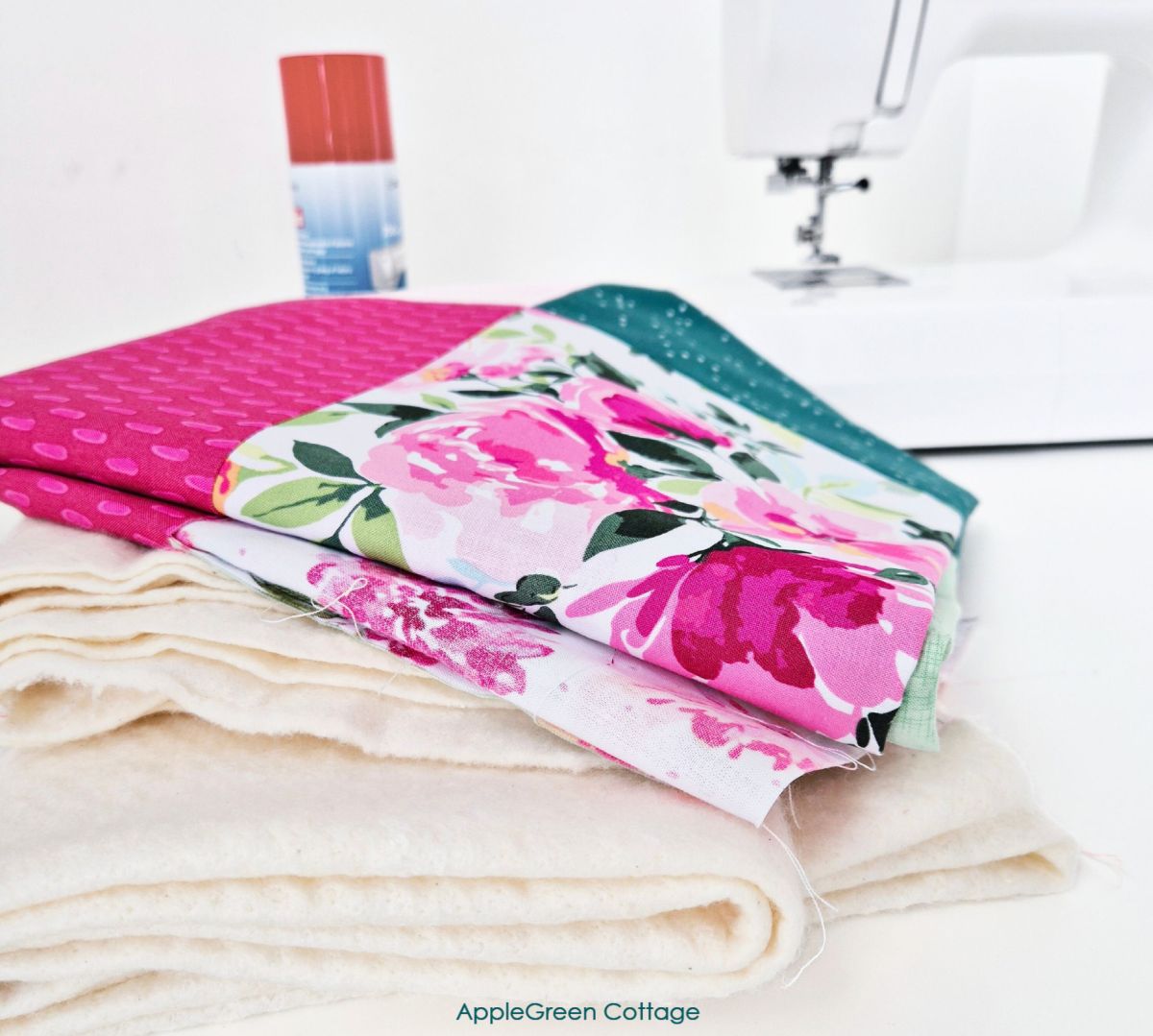 Quilt basting materials including batting, patchwork quilt top, and basting spray near a sewing machine.