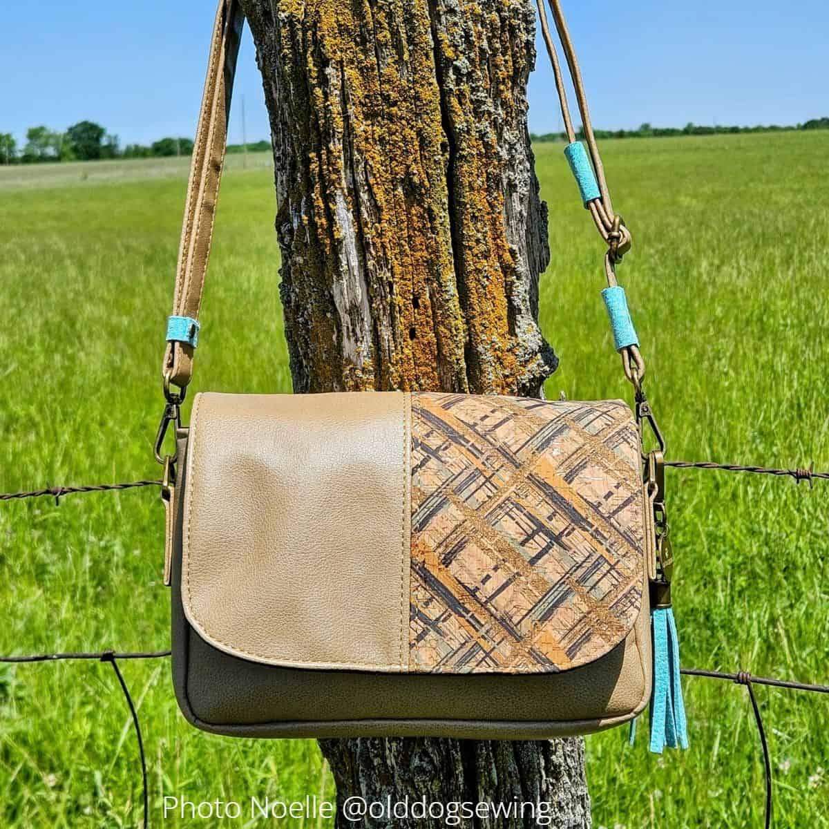 A small handmade saddle bag with a flap, sewn with brown cork and vinyl for the exterior and Ottertex for the interior, hanging on a rustic fence post.