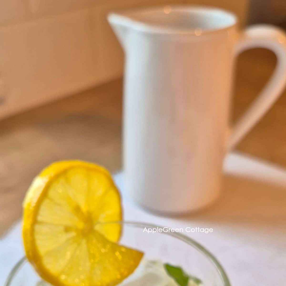 Glass of mint tea with lemon slice beside a white ceramic pitcher on the table.