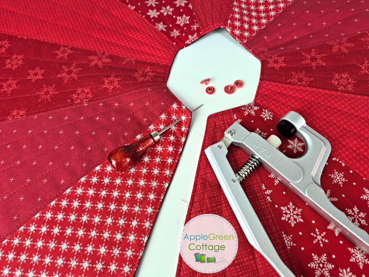 Attaching red snaps with pliers to a quilted Christmas tree skirt sewing project.
