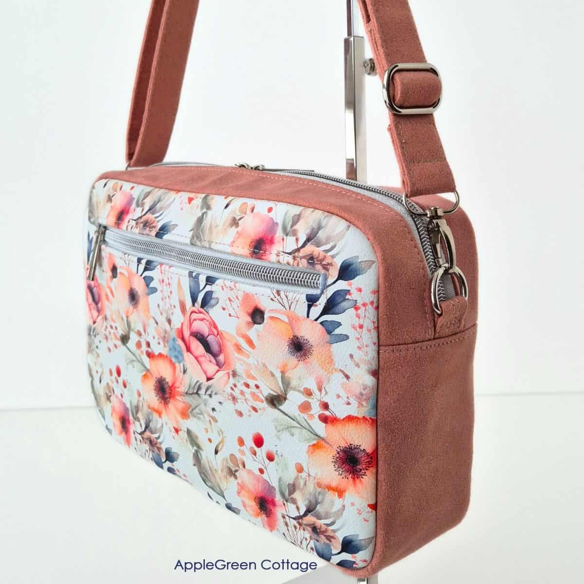 A finished boxy crossbody bag with floral front pocket, double zipper top, and adjustable strap in faux suede.