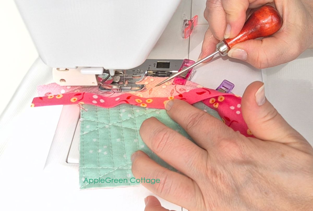 Guiding bias tape with awl near presser foot while stitching curved quilt edge.