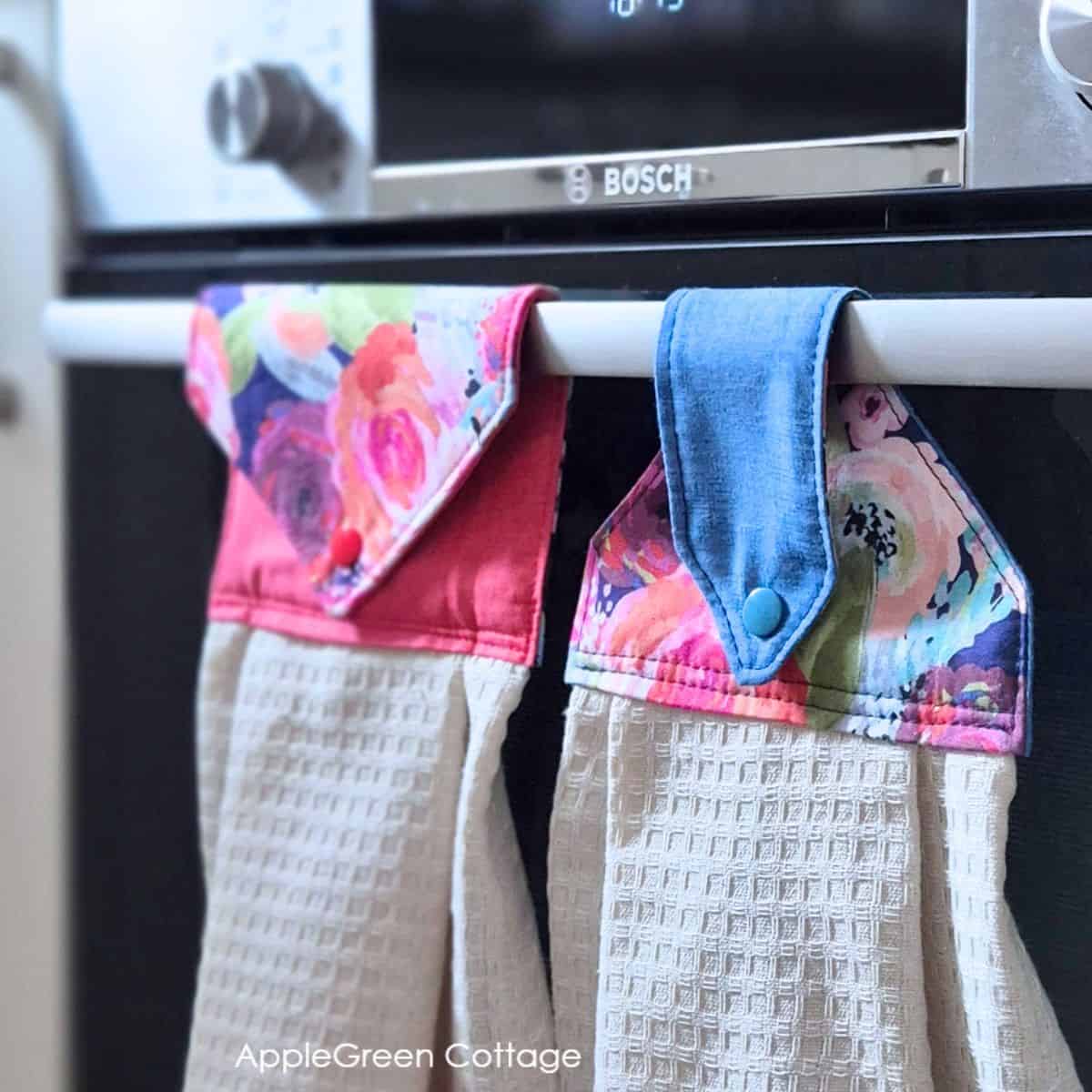 Two finished hanging kitchen towels with floral snap tab toppers on an oven handle.